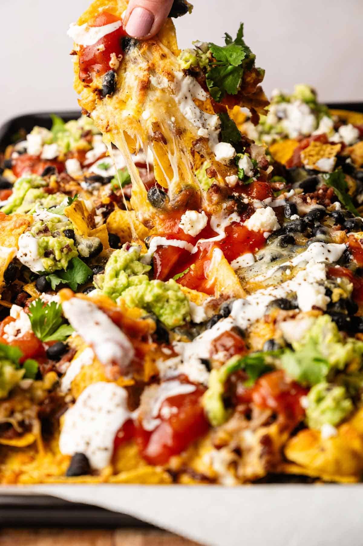 A hand lifts a cheesy nacho chip from a tray of Sheet Pan Nachos topped with guacamole, black beans, tomatoes, sour cream, and cilantro.