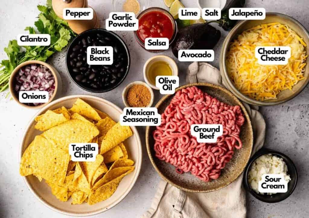 Various taco ingredients—including ground beef, tortilla chips, black beans, cheese, salsa, avocado, sour cream, onions, cilantro, and seasonings—are arranged on a table for assembling delicious Sheet Pan Nachos.
