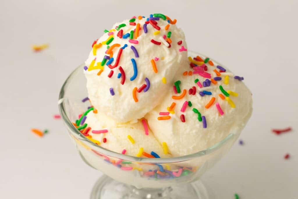 Glass dish with two scoops of Vanilla Ninja Creami Ice Cream topped with colorful rainbow sprinkles, set against a plain white background.