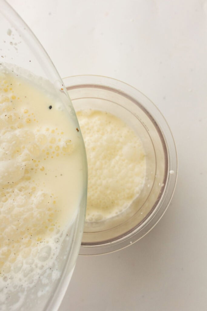 A close-up of frothy Vanilla Ninja Creami Ice Cream being poured from a glass bowl into a clear plastic cup on a white surface.