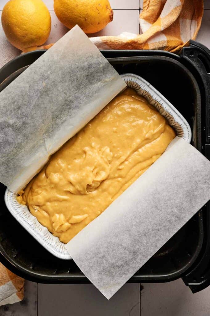 Unbaked lemon cake batter in a lined loaf pan set inside the air fryer, fresh lemons nearby