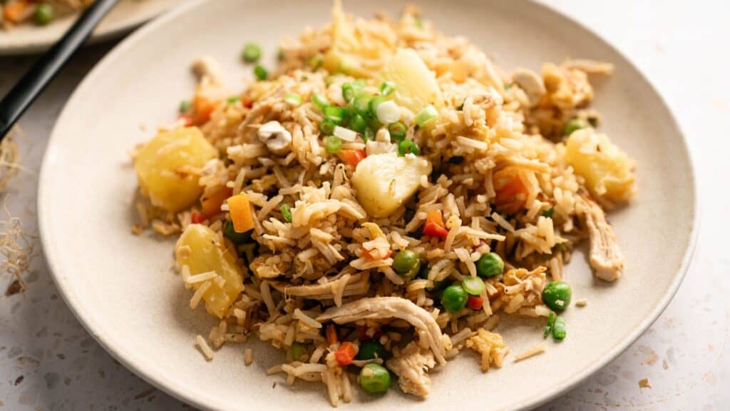 A plate of fried rice with pieces of pineapple, peas, carrots, shredded chicken, and green onions.