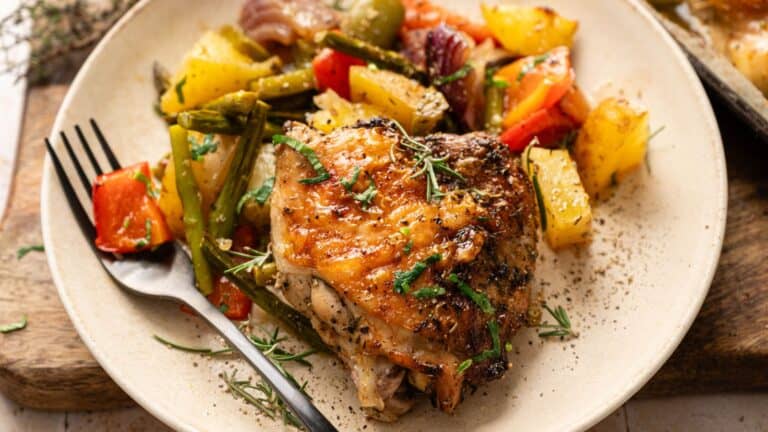 A plate with roasted chicken thigh, assorted roasted vegetables including potatoes, peppers, onions, and green beans, garnished with herbs. A black fork rests on the plate.