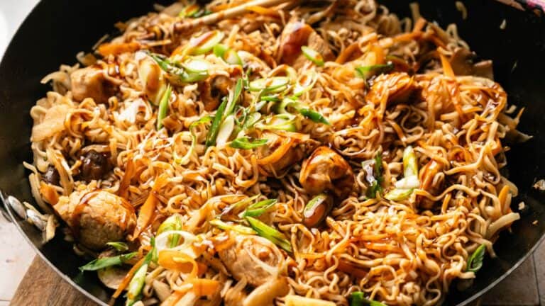 A pan filled with stir-fried noodles, vegetables, chicken pieces, and garnished with sliced green onions and sauce.
