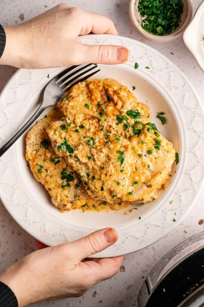 Hands holding a plate of pork chops topped with creamy sauce and parsley