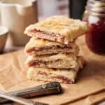 A stack of four sourdough pop tarts with a fruit jam filling and white icing drizzle, resting on brown parchment paper with two silver forks, a jar of jam, and a white pitcher in the background.