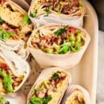 Six air fryer breakfast burritos wrapped in white paper, filled with eggs, sausage, and green salsa, topped with sliced green onions and cilantro in a cream-colored baking dish.