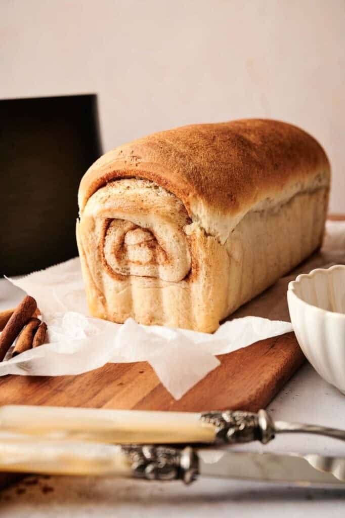 A whole loaf of Air Fryer Cinnamon Swirl Bread with a golden-brown top and a visible cinnamon spiral on the end, resting on a wooden board with vintage knives, cinnamon sticks, and a white fluted bowl.