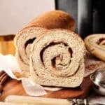 A sliced loaf of Air Fryer Cinnamon Swirl Bread revealing a tight cinnamon-sugar spiral, served on a wooden board with two vintage knives, cinnamon sticks, and a dusting of ground cinnamon.