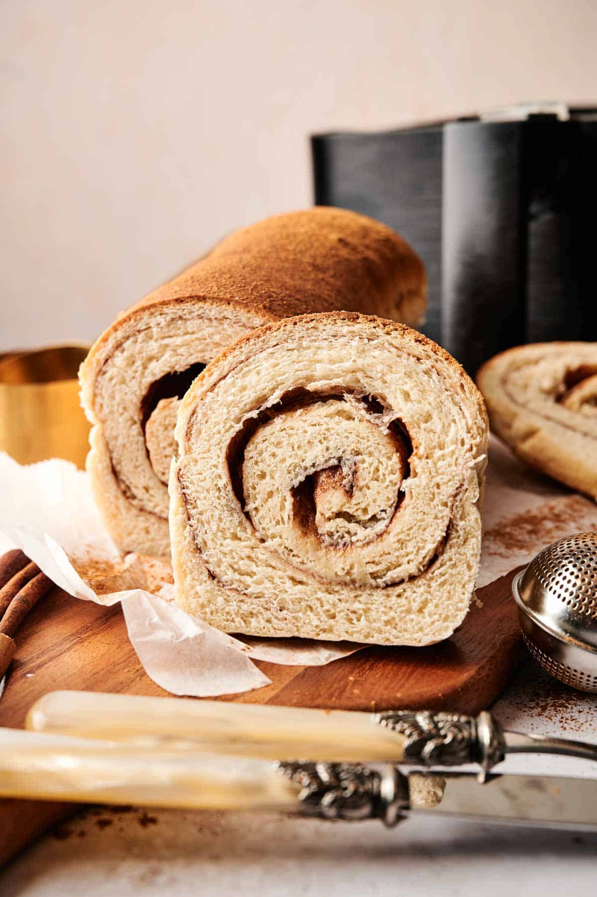 A sliced loaf of Air Fryer Cinnamon Swirl Bread revealing a tight cinnamon-sugar spiral, served on a wooden board with two vintage knives, cinnamon sticks, and a dusting of ground cinnamon.