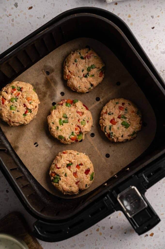 Five uncooked salmon croquette patties arranged in a single layer on parchment paper in a black air fryer basket, ready to cook.