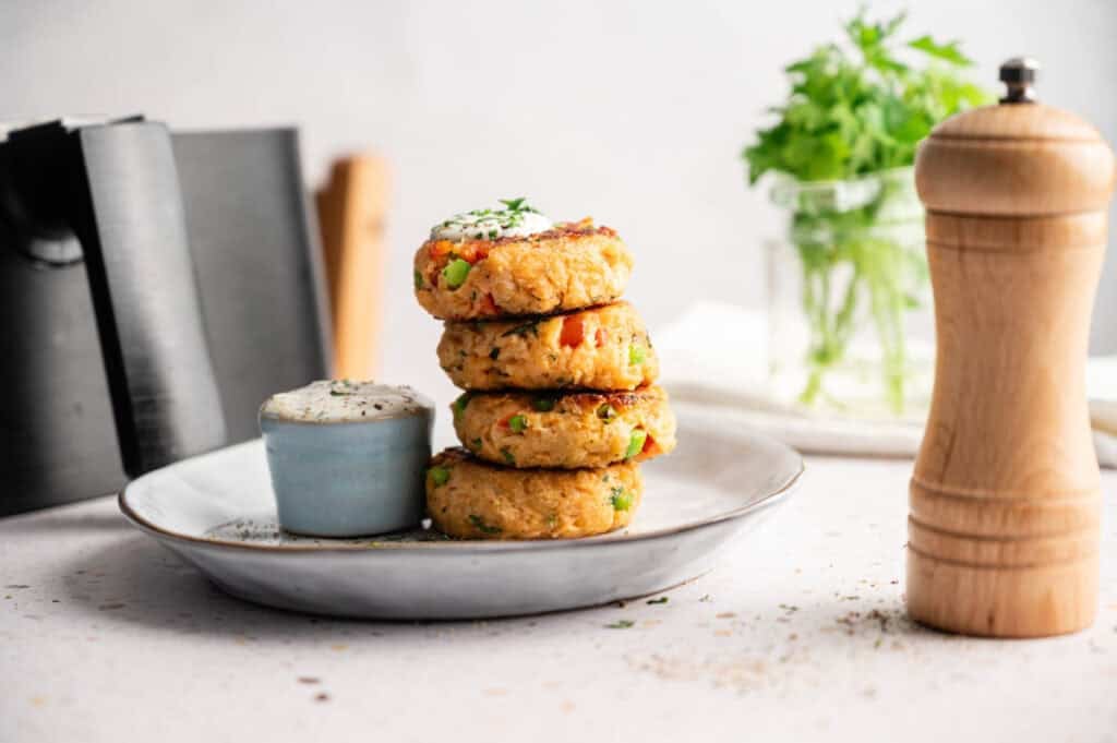 Four air fryer salmon croquettes stacked on a white plate with a small bowl of creamy dipping sauce, garnished with herbs, with a pepper grinder, knife block, and a jar of fresh herbs in the background.