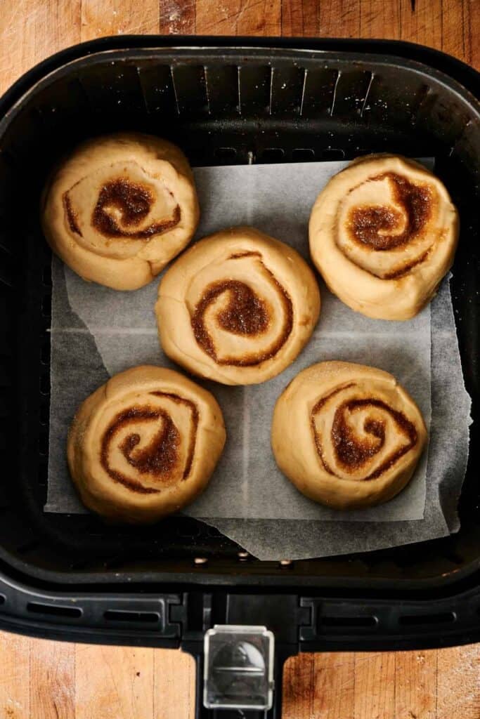 Five unbaked sourdough cinnamon rolls with cinnamon sugar swirls arranged on parchment paper inside a black air fryer basket