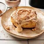 A single air fryer sourdough cinnamon roll covered in dripping cream cheese frosting and dusted with ground cinnamon, served on a rustic beige plate with a small bowl of cinnamon, a sifter, and silver forks in the background.