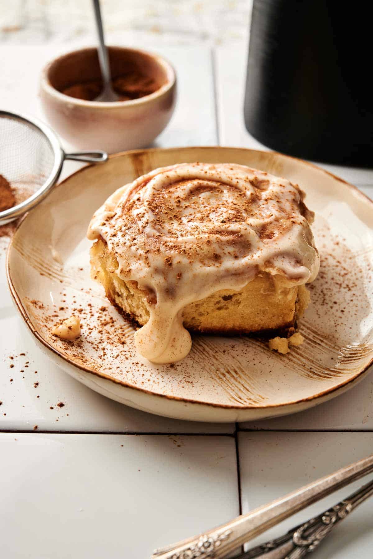 A single air fryer sourdough cinnamon roll covered in dripping cream cheese frosting and dusted with ground cinnamon, served on a rustic beige plate with a small bowl of cinnamon, a sifter, and silver forks in the background.