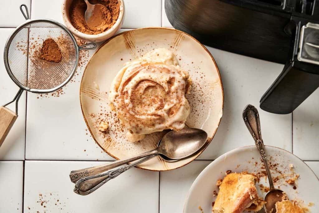 An overhead shot of one frosted air fryer sourdough cinnamon roll dusted with cinnamon on a rustic plate, served with two silver spoons and a second partial serving, with a sifter, a bowl of ground cinnamon, and the corner of a black air fryer in the background.