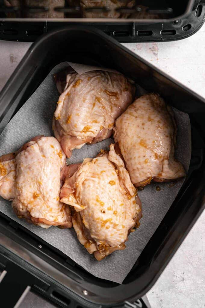 Four marinated raw chicken thighs arranged skin-side down on parchment paper in a black air fryer basket, viewed from above.