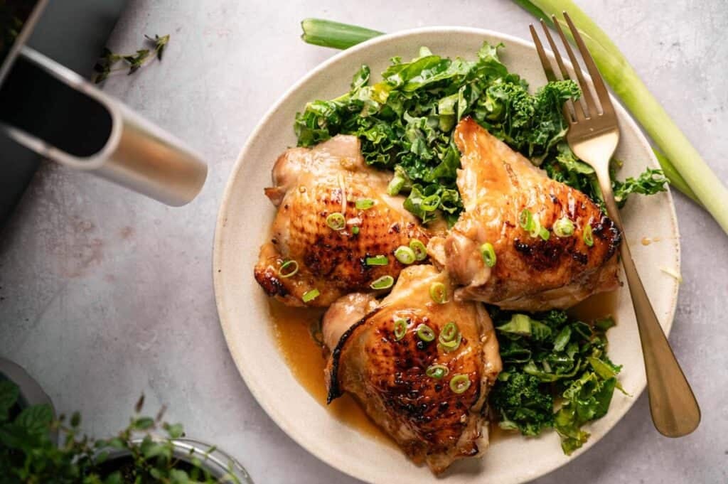 Three Air Fryer Sticky Chicken Thighs with charred, glazed skin topped with sliced green onions, served on a beige plate over a bed of saut&eacute;ed kale with a gold fork, an air fryer basket, and fresh herbs in the background.