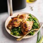 Three Air Fryer Sticky Chicken Thighs with a glossy, charred glaze and a topping of sliced green onions, served on a bed of sautéed kale with a gold fork, positioned next to a black air fryer, a bottle of olive oil, and fresh green onion stalks.
