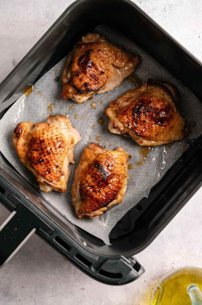 Four Air Fryer Sticky Chicken Thighs with browned, glistening skin resting on a sheet of parchment paper inside a black air fryer basket, with a bottle of oil visible in the bottom corner.