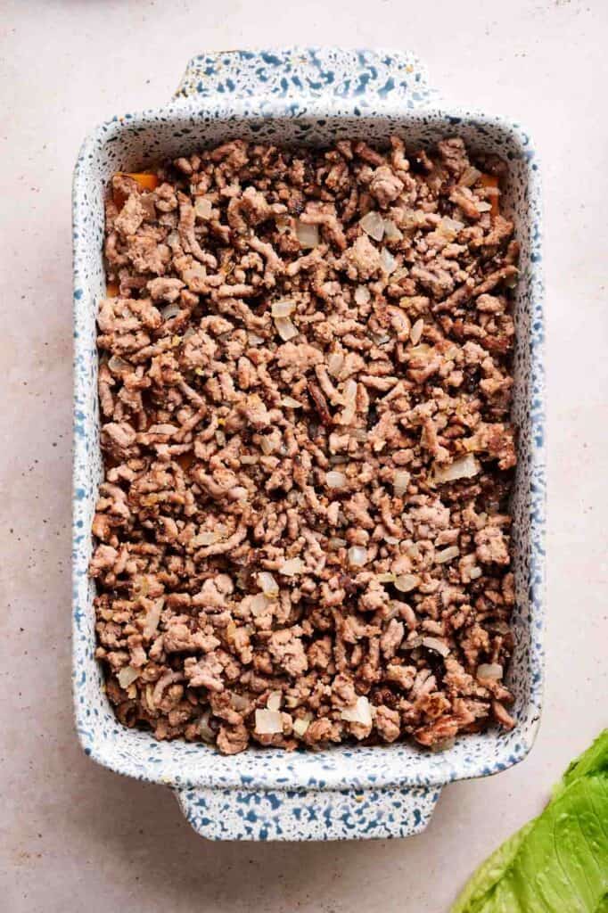Browned ground beef with diced onions spread over slider bun bottoms in a rectangular baking dish for Big Mac sliders.