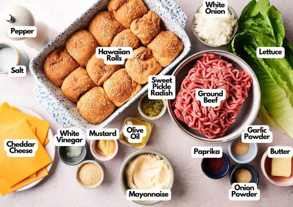 An ingredient spread for Air Fryer Big Mac Sliders featuring twelve Hawaiian rolls, raw ground beef, cheddar cheese, and fresh lettuce. Bowls of white onion, pickles, mayonnaise, and various seasonings are arranged on a pink speckled surface.