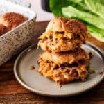 Two stacked Air Fryer Big Mac Sliders on a grey plate, featuring toasted sesame seed buns, melted cheddar cheese, and ground beef. Fresh romaine lettuce and a blue-and-white speckled baking dish are visible in the background on a dark wooden surface.