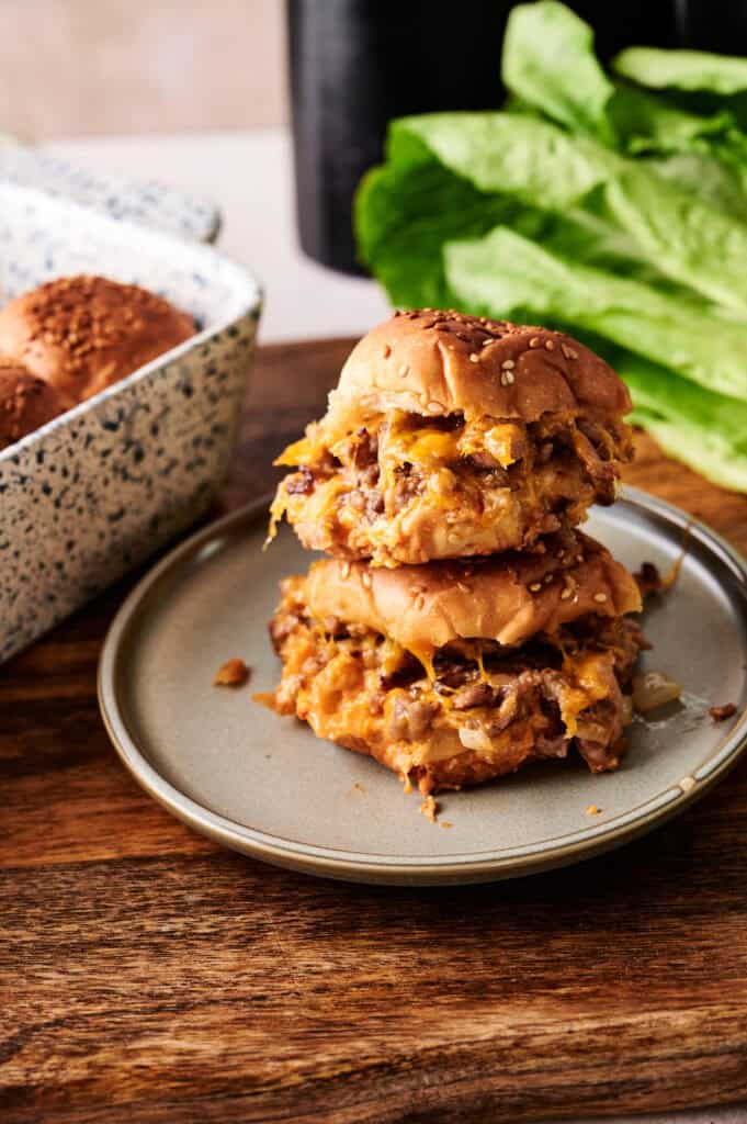 Two stacked Air Fryer Big Mac Sliders on a grey plate, featuring toasted sesame seed buns, melted cheddar cheese, and ground beef. Fresh romaine lettuce and a blue-and-white speckled baking dish are visible in the background on a dark wooden surface.