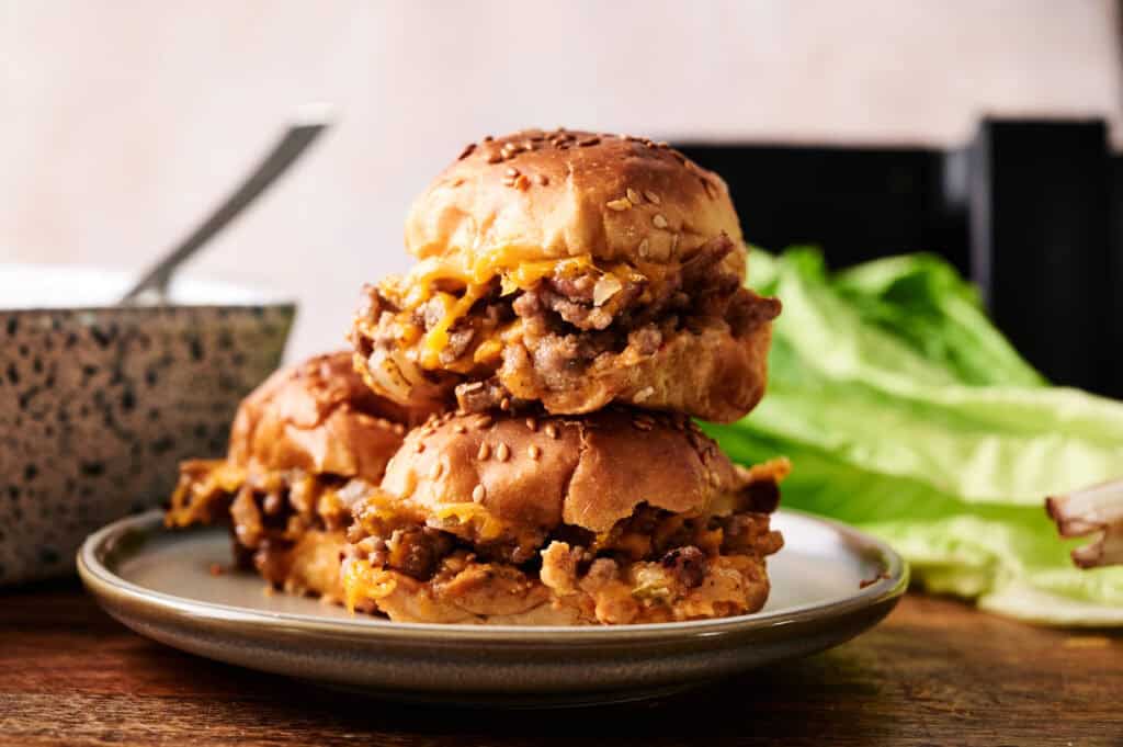 Three Air Fryer Big Mac Sliders stacked on a grey plate, featuring toasted sesame seed buns, melted cheese, and seasoned ground beef. A head of romaine lettuce and a speckled bowl with a spoon are visible in the background on a wooden surface.