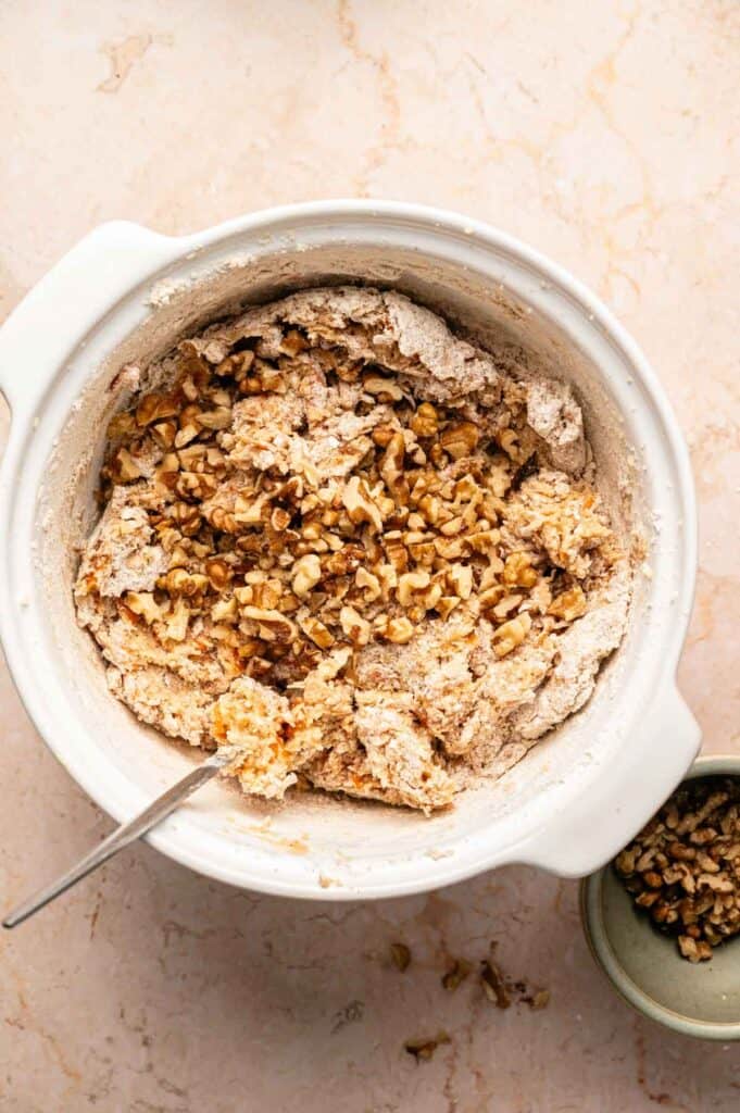 Cookie dough being mixed with dry ingredients and walnuts in a white bowl
