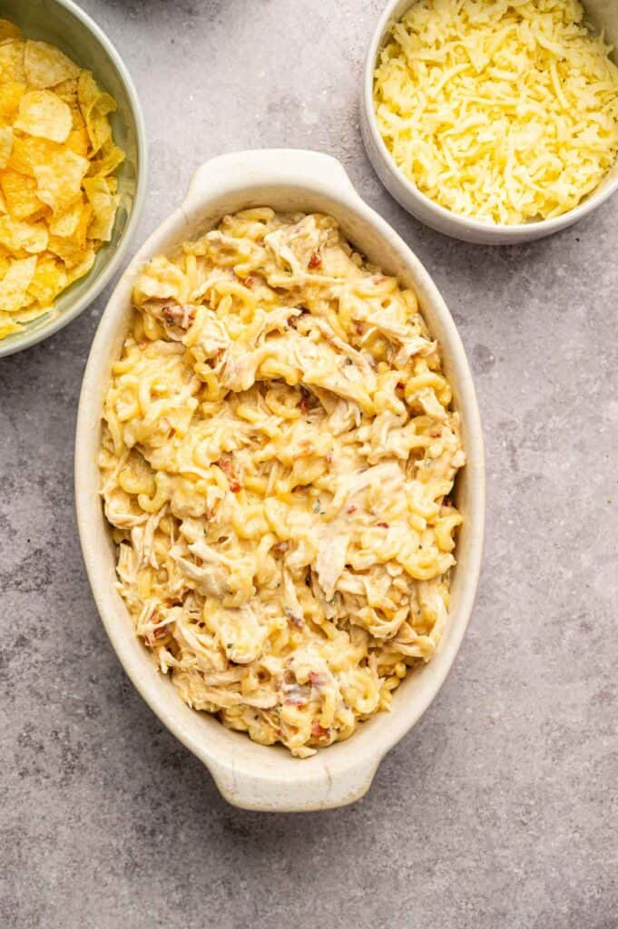 Creamy pasta casserole in a ceramic dish next to bowls of shredded cheese and crushed potato chips
