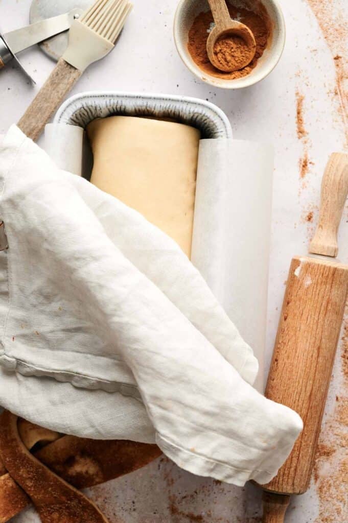 Rolled cinnamon swirl bread dough placed in a parchment-lined loaf pan, covered with a linen cloth to rise, with a rolling pin and bowl of cinnamon nearby.