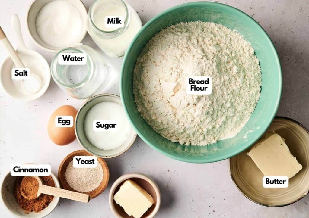 An overhead, flat-lay shot of pre-measured ingredients for Air Fryer Cinnamon Swirl Bread, featuring a large teal bowl of bread flour, a glass bottle of milk, two bowls of sugar, and two portions of butter, accompanied by a single egg, small bowls of salt, yeast, and water, and a bowl of cinnamon with a wooden spoon, all set against a light grey textured background.