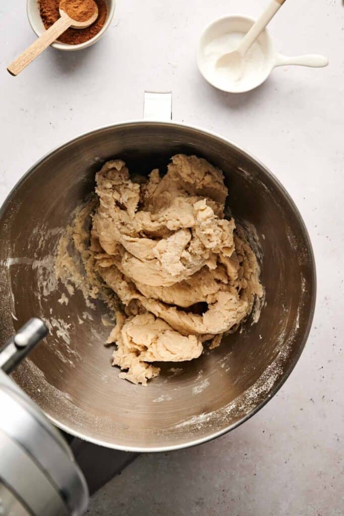 Cinnamon swirl bread dough mixed in a stand mixer bowl, with bowls of cinnamon and sugar in the background.