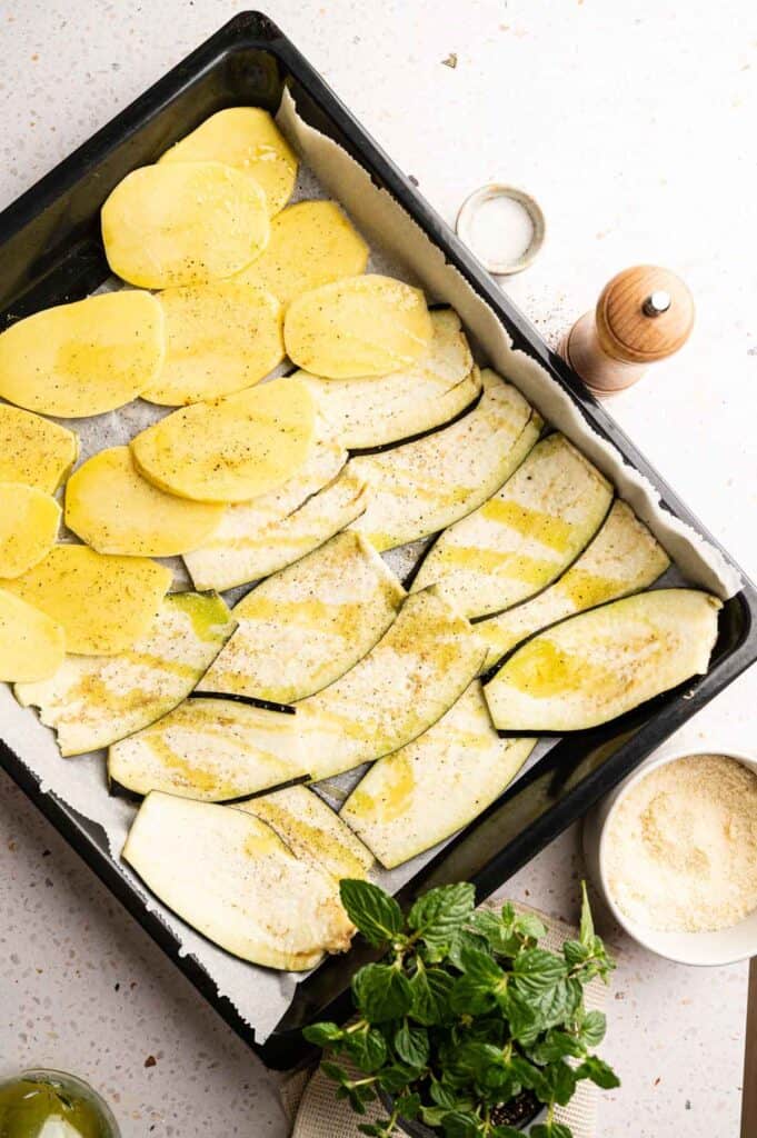 Raw potato and eggplant slices arranged on a tray with olive oil and seasoning