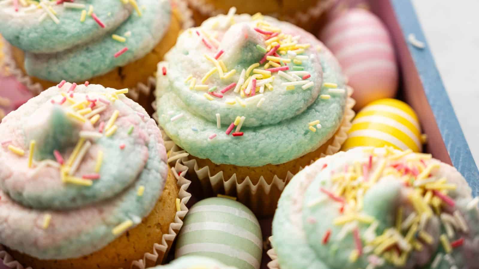 Cupcakes with pastel blue and pink frosting, topped with sprinkles, are arranged next to decorated pastel Easter eggs in a wooden tray.