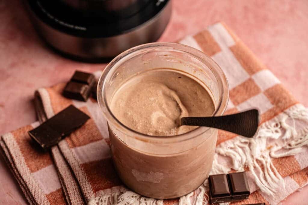 A full pint of Ninja Creami chocolate ice cream in a clear plastic container with a black spoon resting inside, garnished with three separate pieces of dark chocolate bars on a pink and orange checkered towel, with the base of the Ninja Creami machine visible in the background.