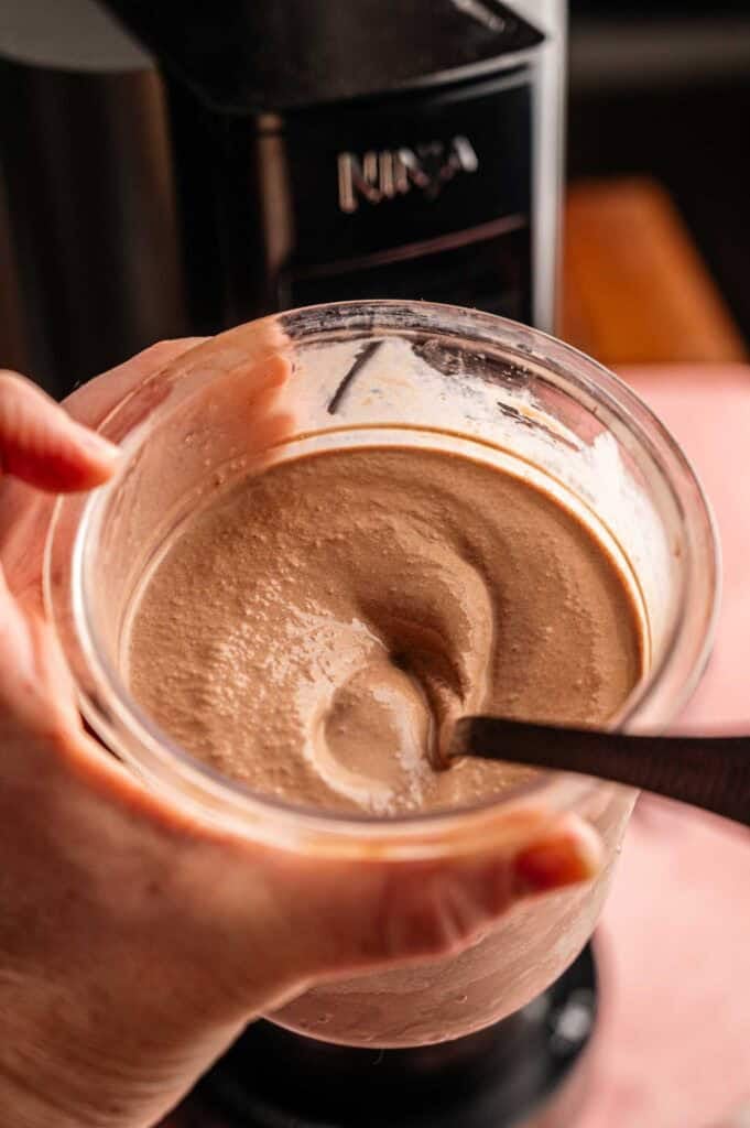 A hand holding a full pint of freshly spun Ninja Creami chocolate ice cream in a clear plastic container with a black spoon creating a thick swirl, set against a pink surface with a black Ninja Creami machine in the background.