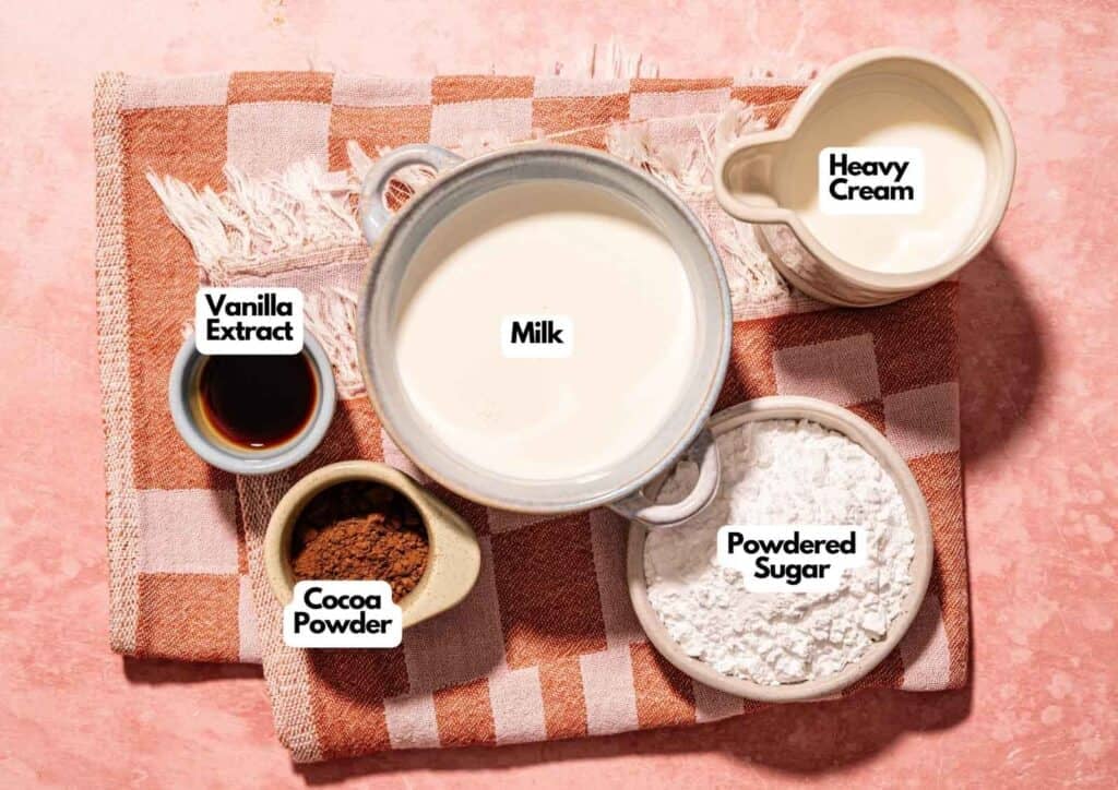 
Gemini said
An overhead, processed shot of ingredients for Ninja Creami Chocolate Ice Cream arranged on a pink and orange checkered towel: one bowl of milk, one small pitcher of heavy cream, a bowl of powdered sugar, a small dish of cocoa powder, and a small ramekin of vanilla extract. Each ingredient is clearly labeled with a white text bubble.