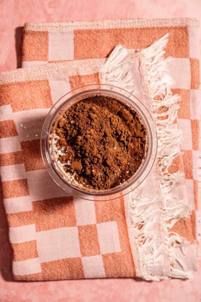 Overhead view of a glass bowl of cocoa powder on a checkered kitchen towel &mdash; a key ingredient for homemade chocolate ice cream.