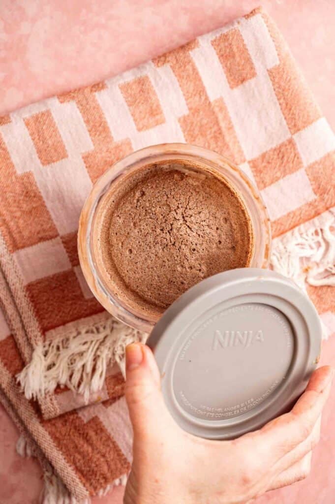 A hand lifting a gray Ninja-branded lid from a full pint of Ninja Creami chocolate ice cream, showing the frozen, textured surface of the chocolate base inside the clear container, set against a pink and orange checkered towel.
