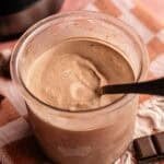 A full pint of Ninja Creami chocolate ice cream in a clear container with a black spoon, garnished with four pieces of dark chocolate bars arranged on a pink and orange checkered towel, with a Ninja Creami machine in the background.