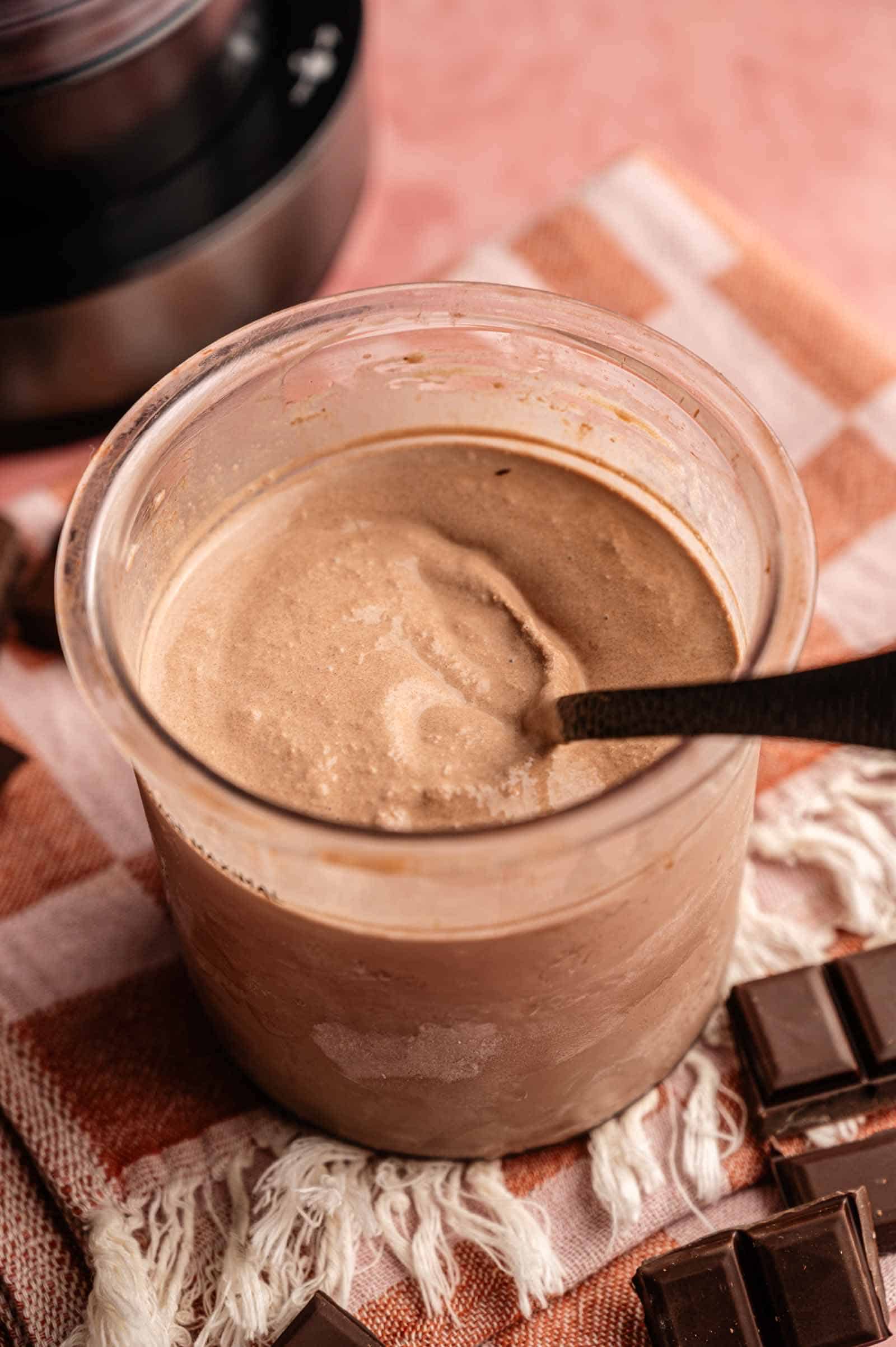A full pint of Ninja Creami chocolate ice cream in a clear container with a black spoon, garnished with four pieces of dark chocolate bars arranged on a pink and orange checkered towel, with a Ninja Creami machine in the background.