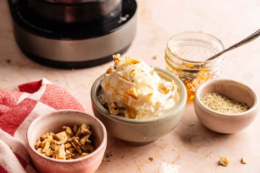 Three bowls of Ninja Creami frozen yogurt topped with granola and honey, served on a grey surface with a whole mango, a half pineapple, and a small side bowl of extra granola.