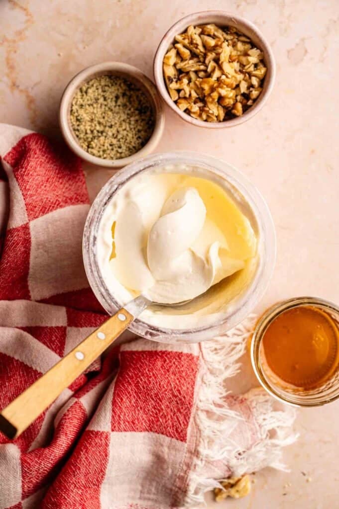 One pint of freshly spun Ninja Creami frozen yogurt in its clear container, served with a wooden-handled spoon and side bowls of chopped walnuts, hemp seeds, a jar of honey, and a red checkered napkin.