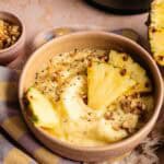 A Ninja Creami smoothie bowl topped with three pineapple slices and crushed walnuts, served with side bowls of walnuts and hemp seeds, and a half pineapple in the background.