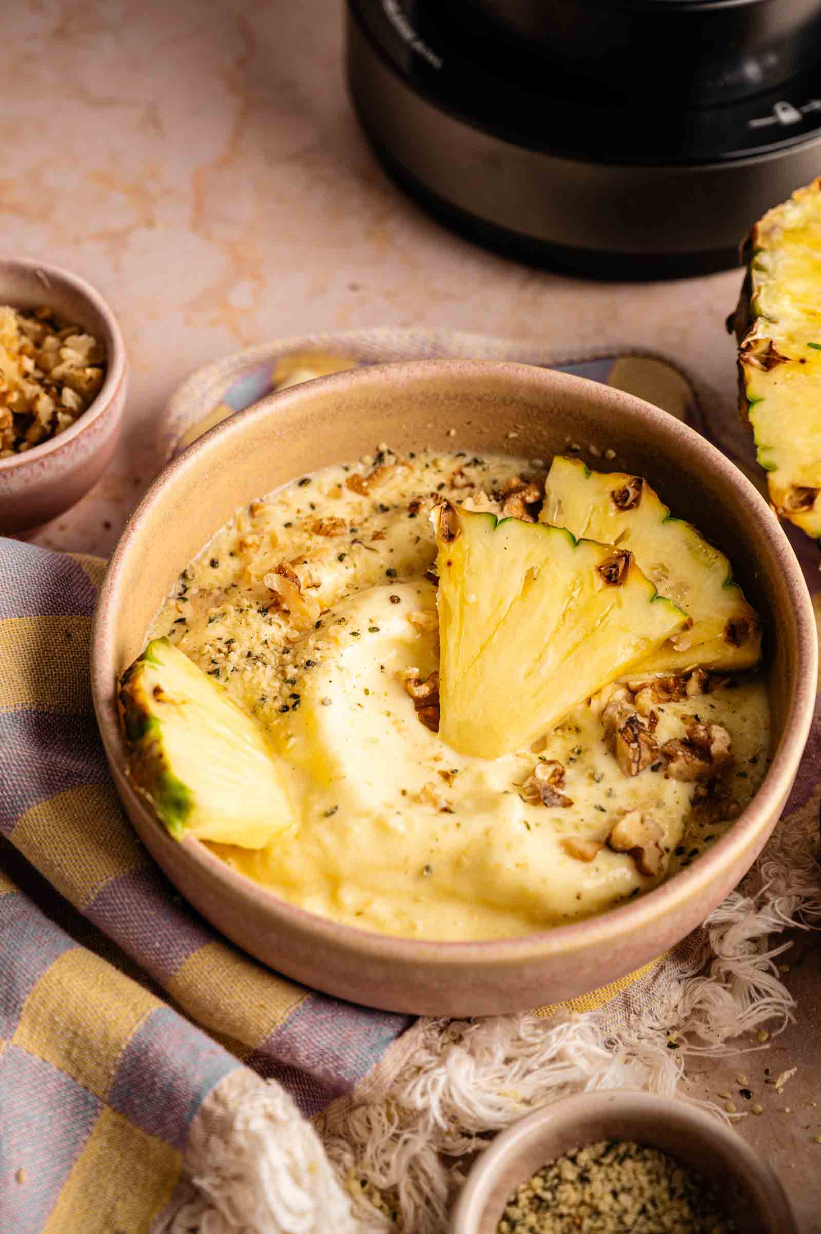 A Ninja Creami smoothie bowl topped with three pineapple slices and crushed walnuts, served with side bowls of walnuts and hemp seeds, and a half pineapple in the background.