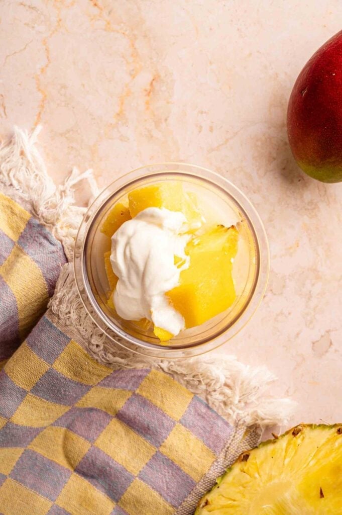 Process shot of pineapple chunks topped with yogurt in a glass container, prepared as a base for a Ninja Creami smoothie bowl, on a light marble surface with fresh mango and pineapple nearby.