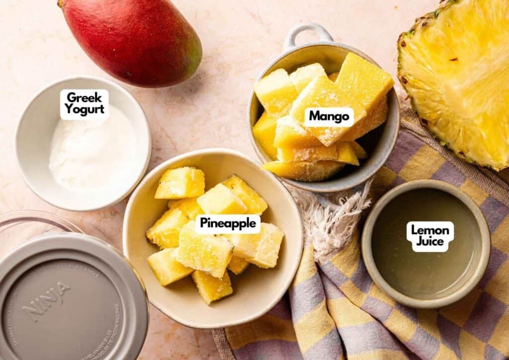 Ingredients for a Ninja Creami Smoothie Bowl including bowls of frozen mango chunks, frozen pineapple chunks, Greek yogurt, and lemon juice, with a whole mango, a half pineapple, and a Ninja Creami lid on the side.
