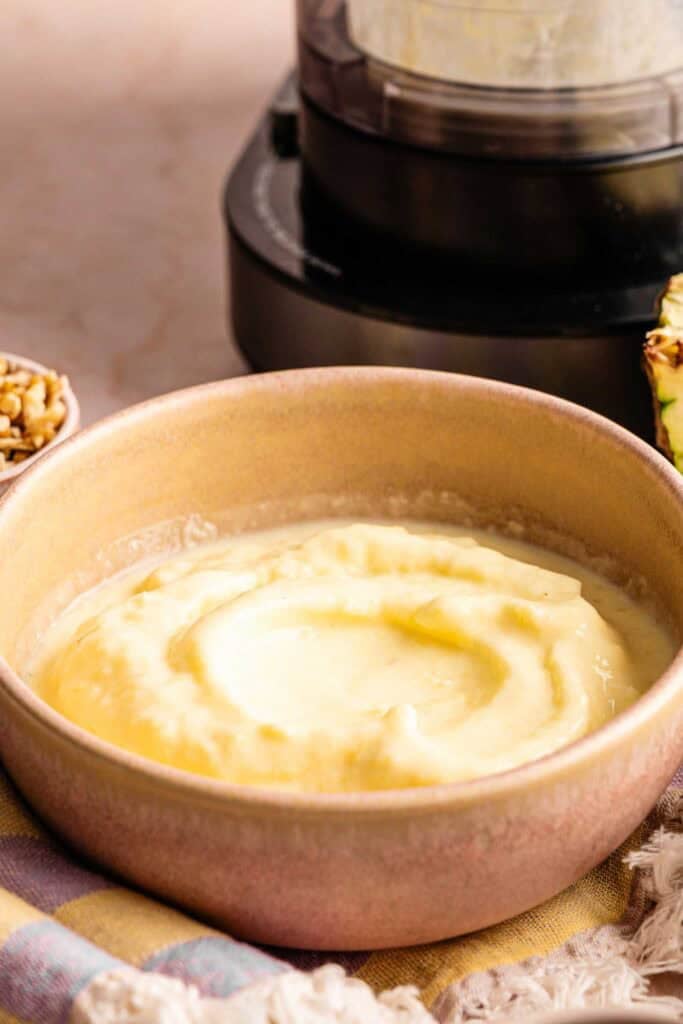 A Ninja Creami smoothie bowl topped with three pineapple slices and crushed walnuts, served with a side bowl of walnuts, a half pineapple, and a whole mango in the background.